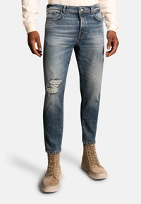 Blue denim jeans with a slim fit, featuring a distressed knee detail and subtle fading, paired with tan lace-up boots.