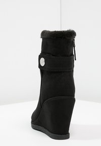 Bottes compensées noires avec une douce bordure en fausse fourrure, fermeture éclair et détail en métal argenté circulaire. Fabriquées en matériau semblable au daim avec une semelle texturée.