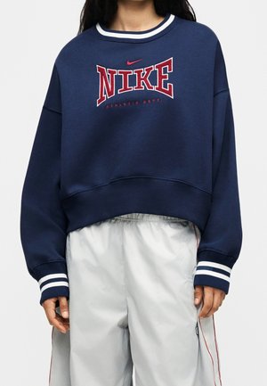 Person iført en mørkeblå Nike sweatshirt med rød logo og hvide stribede manchetter, kombineret med lysegrå løse bukser med røde sidelister.