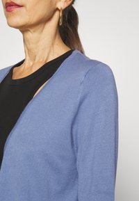 Cardigan bleu clair en maille douce, présentant un col en V et des manches montées, porté sur un haut noir. Boucles d'oreilles anneaux dorés visibles.