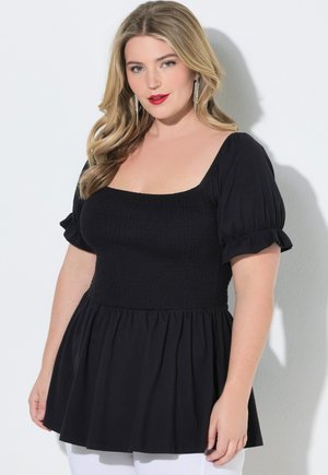 Kobieta o długich blond włosach nosi czarną bluzkę z kwadratowym dekoltem i bufiastymi rękawami oraz białe spodnie, stojąc przy białej ścianie.