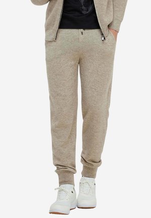 Beige gestrickte Jogginghose mit elastischen Bündchen, getragen mit weißen Sneakern, kombiniert mit einer passenden Zip-Up-Jacke und schwarzem Shirt.