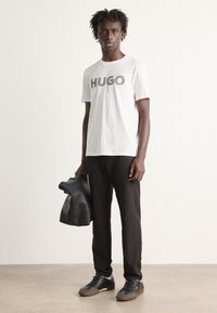 Hvit bomullstrøye med "HUGO" logo, svarte slim-fit bukser, svart skinnveske, og grå joggesko med brune såler, stående på et teksturert gulv.