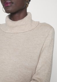 Maglione a collo alto beige realizzato in morbido materiale a maglia con colletto a coste. Presenta una texture liscia e una vestibilità rilassata.