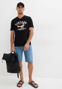 T-shirt noir à manches courtes avec impression vintage, shorts en denim bleu clair, sac à dos noir et sandales grises ; tenue décontractée présentée.