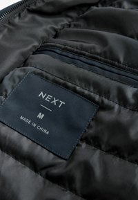 Interiorul unei jachete negre căptușite, cu o etichetă din material navy pe care scrie "NEXT," mărimea "M," și "FABRICAT ÎN CHINA." Include o buzunare cu fermoar.