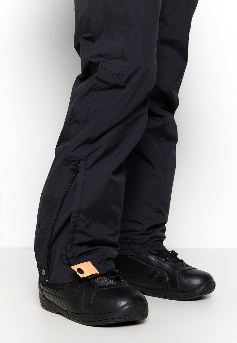 Czarne wodoodporne spodnie z zamkami po bokach, z pomarańczowym akcentem guzikowym, w zestawie z czarnymi teksturowanymi butami sportowymi z gumową podeszwą.
