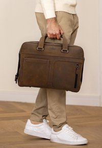 Brun læder aktetaske med to hanke, forlommer og sølv hardware. Båret af en person i lyse bukser og hvide sneakers.