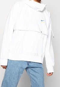Valkoinen huppari, jossa on kenguru-tasku, säädettävä nauhakuulokkeella ja sininen Nike "AIR" -logo. Pehmeä rakenne ja rento istuvuus.