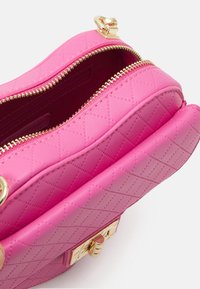 Růžová prošívaná kabelka se dvěma zlatými zipy a kovy. Má texturovaný povrch a strukturovaný tvar. Uvnitř chybí viditelné oddíly.
