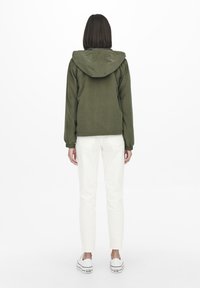 Olivengroene hoodie met elastische manchetten, gedragen over een witte broek en sneakers, gezien vanaf de achterkant tegen een effen achtergrond.