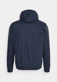 Giacca leggera blu navy con cappuccio, polsini e orlo elastici. Presenta una texture liscia e un design pulito e minimalista.