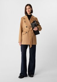 Tan dubbelzijdig jasje met grote zwarte knopen, een strik taille en zijzakken, gecombineerd met donkere flared jeans en zwarte schoenen.