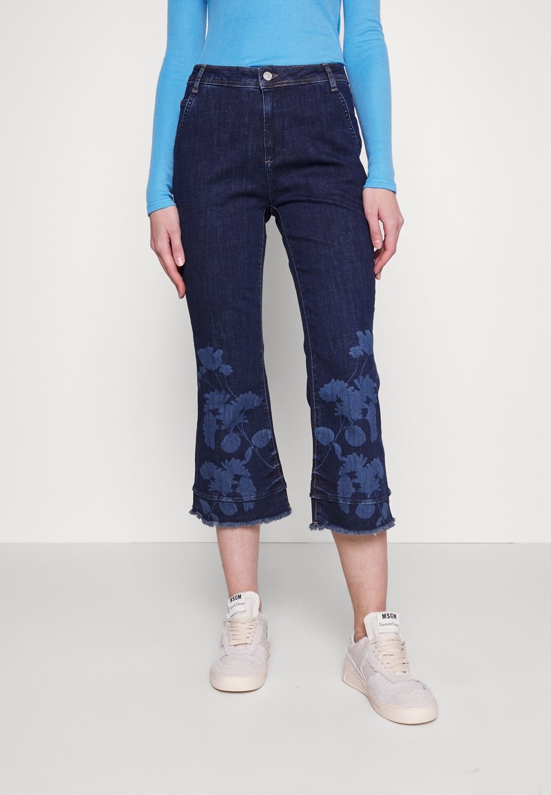 Personne portant un jean court bleu foncé avec des motifs floraux près de l'ourlet, associé à des baskets blanches et un haut à manches longues bleu clair.