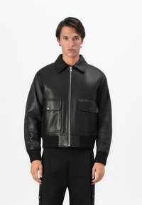 Joven con una chaqueta de cuero negra con grandes bolsillos frontales y una cremallera plateada, de pie frente a un fondo blanco liso.