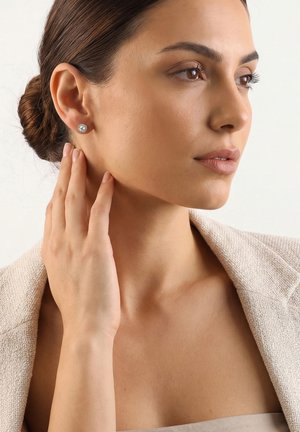 Vrouw met bruin haar in een knot, draagt een beige blazer en diamantsteker oorbellen, raakt voorzichtig haar nek aan terwijl ze opzij kijkt.