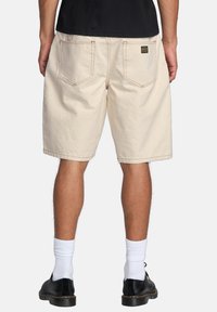Pantaloni corti in cotone beige con vestibilità rilassata, cuciture a vista, due tasche posteriori e un'etichetta nera sulla tasca destra. Indossati con scarpe nere e calze bianche.