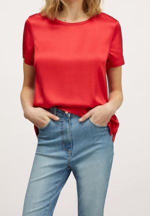 Kobieta w jasnoczerwonej bluzce z krótkim rękawem wpuszczonej w jasnoniebieskie, wysokiego stanu jeansy, z rękami w przednich kieszeniach.