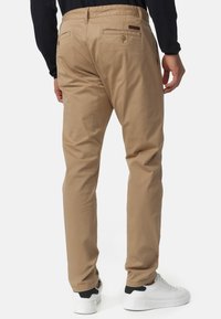 Chinos de mezcla de algodón beige con un diseño ajustado. Presentan dos bolsillos traseros con cierres de solapa y un discreto parche de la marca. Combinados con zapatillas blancas.