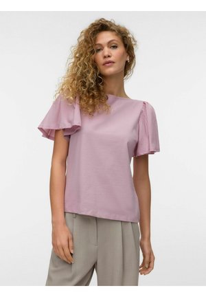 Femme aux cheveux bouclés portant un chemisier lilas à manches courtes évasées et un pantalon beige taille haute, debout devant un fond blanc.