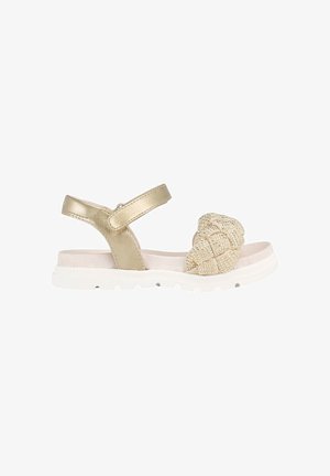 Sandalia beige con suela blanca acolchada, tira metálica alrededor del tobillo y tira frontal trenzada con cuentas.