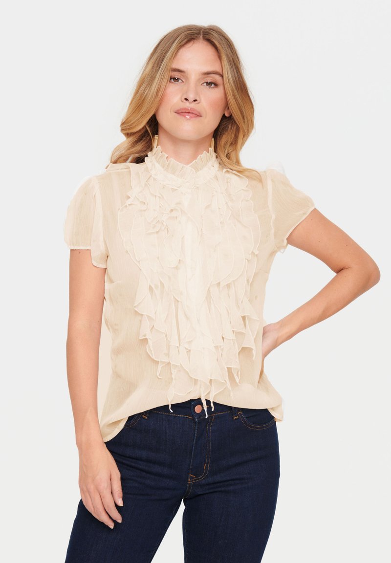 Blouse beige clair avec un devant à volants, col montant et manches bouffantes courtes. Tissu texturé et design fluide, associée à un jean en denim foncé.
