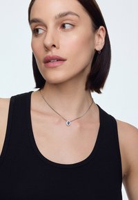 Srebrny naszyjnik z niebieskim kamieniem w centrum, ozdobiony kwiatowym wzorem, na cienkim łańcuszku. Modelka ma na sobie czarny top bez rękawów.