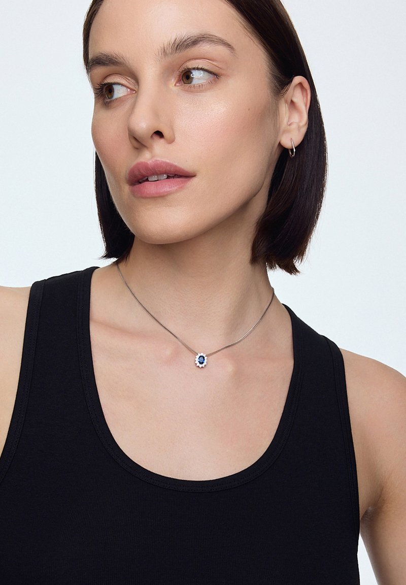 Srebrny naszyjnik z niebieskim kamieniem w centrum, ozdobiony kwiatowym wzorem, na cienkim łańcuszku. Modelka ma na sobie czarny top bez rękawów.