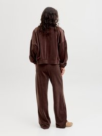 Person with long curly hair wearing loose brown velour tracksuit and beige sneakers, standing facing away on plain white background.