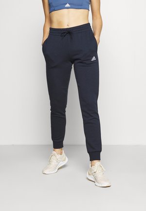 Kobieta ubrana w granatowe spodnie dresowe Adidas z elastycznymi mankietami i białe trampki, stojąca na tle jednolitego jasnego tła.