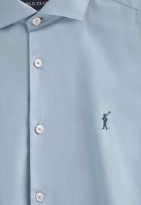 Camicia azzurro chiaro realizzata in tessuto liscio, con colletto classico, bottoni bianchi e una piccola figura ricamata sul petto sinistro.