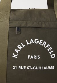 Zöld texturált vászontáska fehér nyomtatott logóval: "KARL LAGERFELD PARIS 21 RUE ST-GUILLAUME", és tartós, állítható füllel.