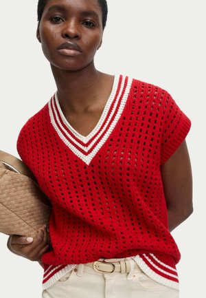 Pull en tricot rouge avec un col en V, des accents de rayures blanches et une texture en treillis ouvert. Tissu léger suspecté, porté avec une pochette beige.