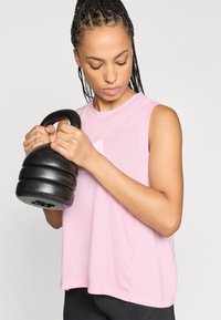 Ροζ αθλητική μπλούζα χωρίς μανίκια με χαλαρή εφαρμογή. Το μοντέλο κρατάει μια μαύρη καμπάνα γυμναστικής, αναδεικνύοντας τονισμένους μύες και συγκέντρωση. Απλός σχεδιασμός, ελάχιστες λεπτομέρειες.