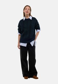 Pull en maille vert foncé avec un motif texturé, superposé à une chemise à col bleu clair, associé à un pantalon large noir et des chaussures brunes.