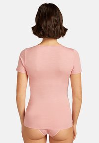 Kvinna med kort brunt hår iklädd en figurnära rosa kortärmad tröja och matchande rosa underkläder, sedd bakifrån mot en enfärgad bakgrund.