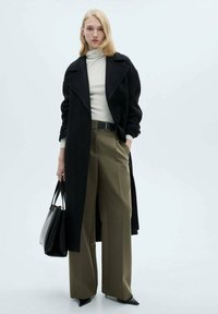 Črn plašč, kremasti turtleneck, olivno široke hlače in koničaste črne pete. Model drži črno torbo in stoji pred svetlo ozadje.