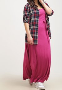 Maxi kjole i lyserød stof med en indsnøring i taljen og en ternet skjorte lagt ovenpå. Har lange ærmer og en afslappet pasform.