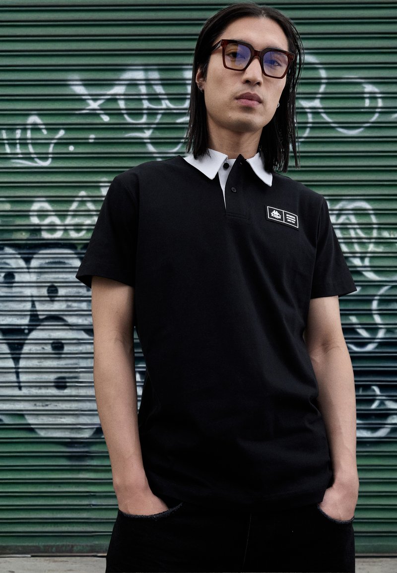 Tricou polo negru cu guler alb și etichetă pe piept. Purtați cu pantaloni negri, pe un fundal verde cu dungi și graffiti.