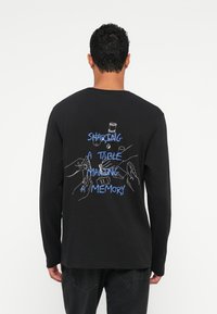 Vīrietis, tērpies melnā garo piedurkņu kreklā ar zilu tekstu "DALOT GALDU, RADOT ATMIŅU" un baltiem zīmējumiem ar rokām un objektiem uz muguras.