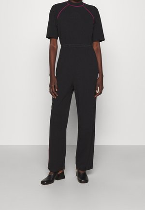 Persona que lleva un mono negro de manga corta con detalles de costuras rosas y zapatos negros de tacón sin cordones, de pie frente a una pared clara y lisa.