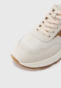 Zapato deportivo beige con material de cuero suave, suela de goma texturizada y cordones planos. Presenta un detalle en marrón contrastante en el lateral.