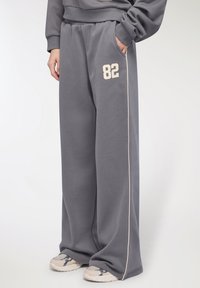 Osoba w luźnych szarych dresach z numerem 82 na udzie, z bocznymi paskami i beżowymi sneakersami na białym tle.