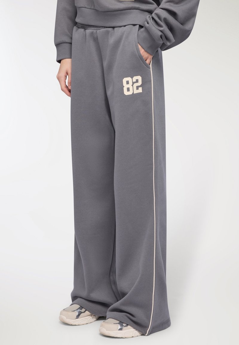 Osoba w luźnych szarych dresach z numerem 82 na udzie, z bocznymi paskami i beżowymi sneakersami na białym tle.