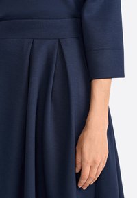 Lumo sininen mekko, jossa on vartalonmyötäinen yläosa ja pleidattu hame. Silkkinen pinta ja jämäkkä vyötärö. Hihat ulottuvat ranteeseen.