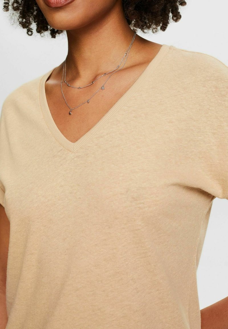Helles beige V-Ausschnitt-T-Shirt mit kurzen Ärmeln, weichem Stoff und lockerer Passform, kombiniert mit einer geschichteten Silberkette.