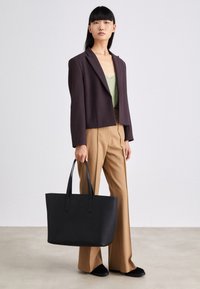 Zwarte shopper met twee handvatten, gladde textuur, rechthoekige vorm, met minimale branding. Gecombineerd met een donker colbert en beige broek.