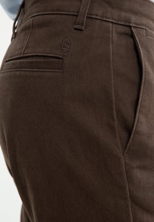 Pantalons marron en tissu texturé, avec une poche arrière portant un logo discret et une coupe ajustée. Coutures soigneusement finies.