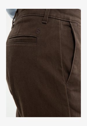 Hnědé kalhoty vyrobené z texturované látky, s praktickou zadní kapsou a decentním logem, v přiléhavém střihu. Úhledně zakončené švy.