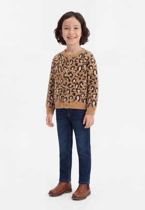Barn smilende, iført en pelsagtig leopardmønstret sweater, mørkeblå jeans og brune ankelstøvler, stående på hvid baggrund.